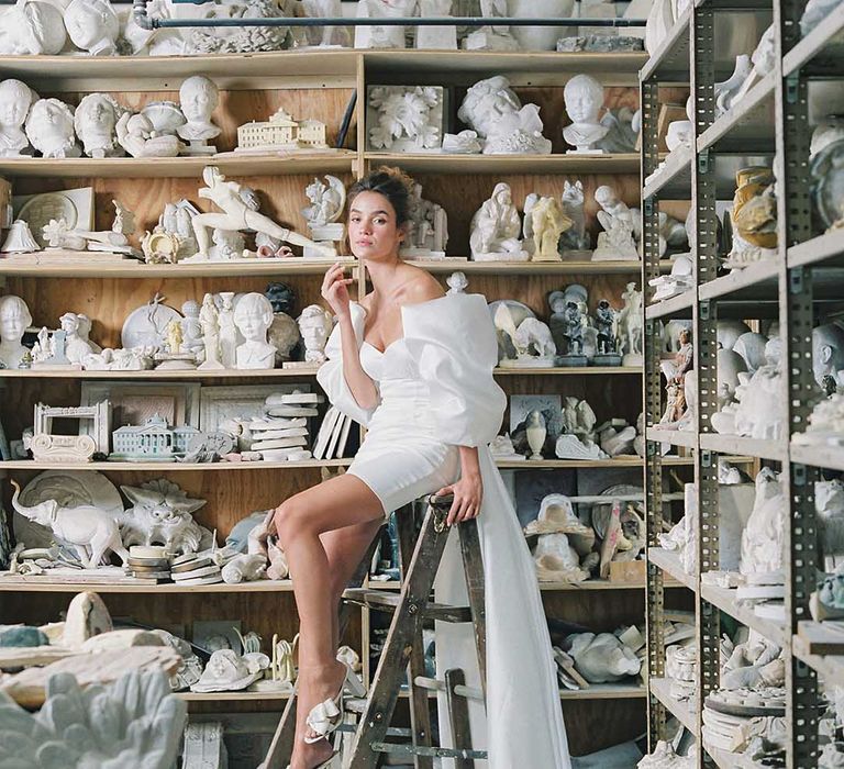 Bride seated on ladder next to figures of heads wearing short wedding dress and mule wedding shoes 