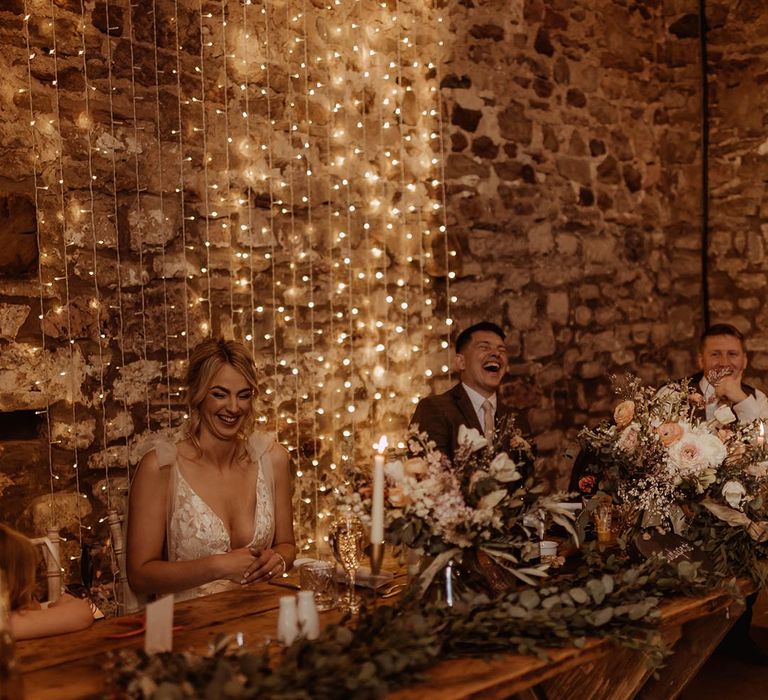 Bride and groom laughing on the wooden top table decorated with foliage runner with white candles and fairy light backdrop 