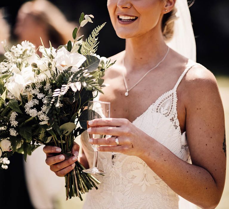 Bride with braided updo wearing lace wedding dress holding small glass of champagne and white flower bridal bouquet 