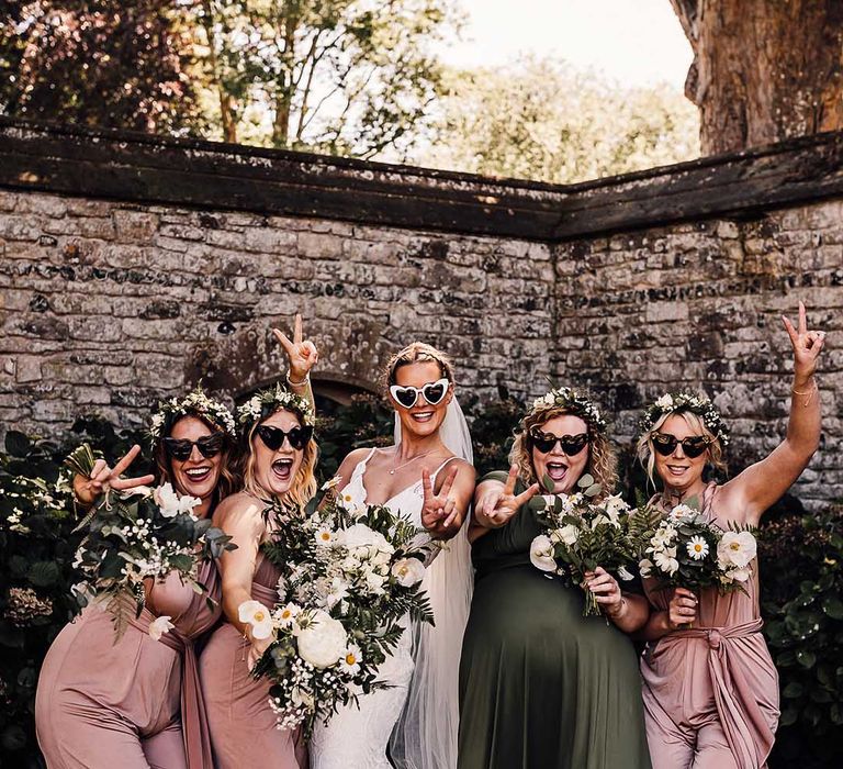 Bridesmaids and bride in retro heart sunglasses throw up peace signs with bridesmaids in pink and green dresses 