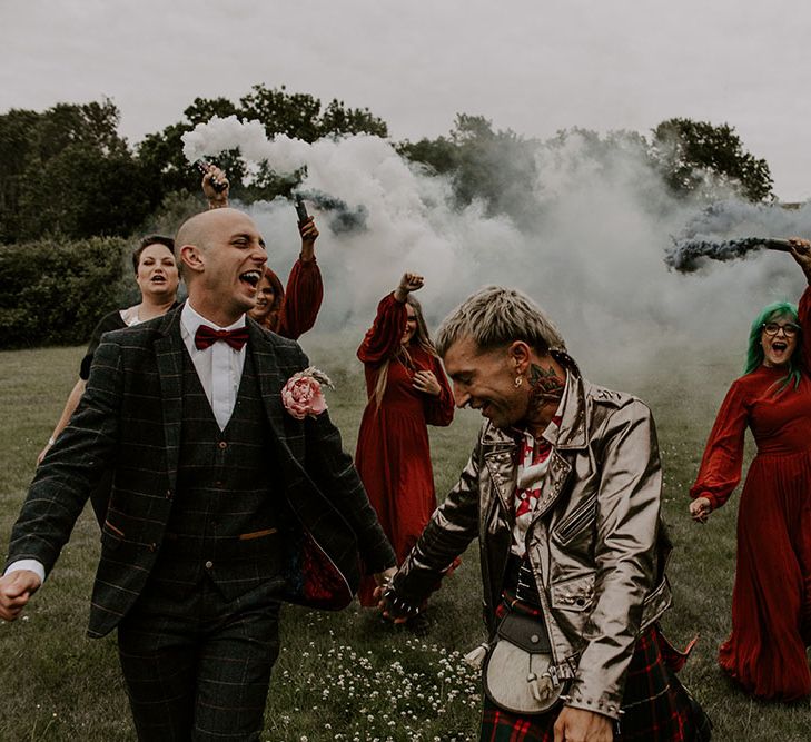 Groom walk through grey smoke bombs beside their bridesmaids who wear long sleeve gowns 