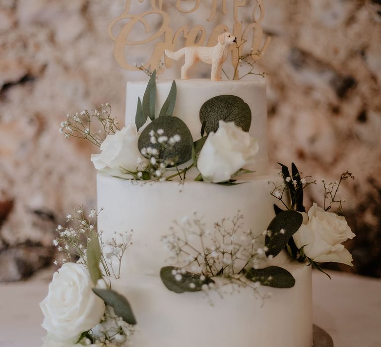 Three tier white wedding cake with white rose decorations and a personalised wooden cake topper with a figurine of their dog 