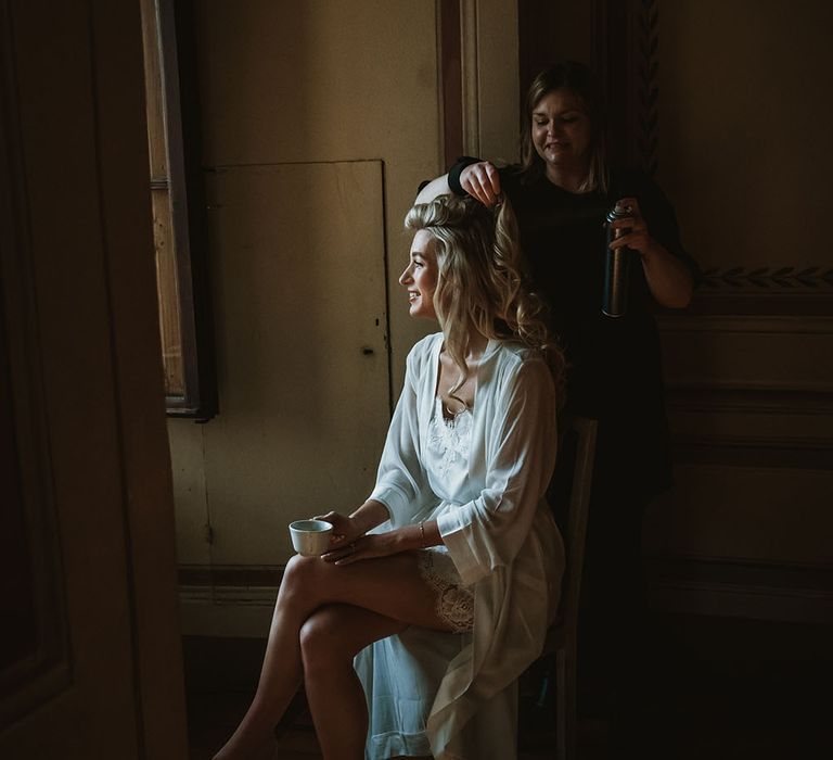 Bride wears satin and lace trim pyjamas on the morning of her wedding day as she has her blonde hair put into loose curls 