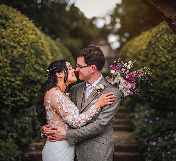 Bride & groom kiss in front of botanical gardens as bride wears sheer long sleeve wedding gown with lace floral embellishment and holds colourful bouquet 