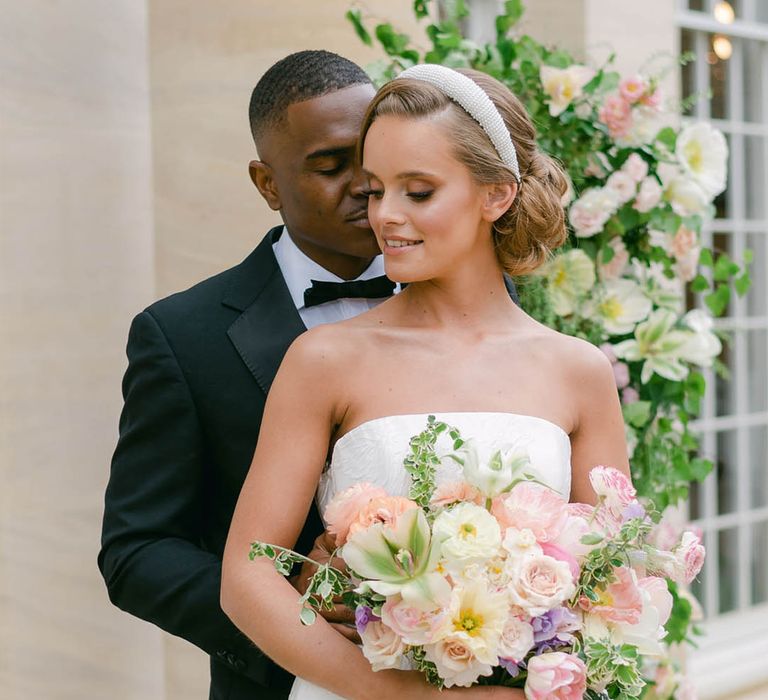 Groom in black tie embraces the bride from behind who wears a strapless wedding dress and beaded headband holding a colourful bouquet with coral pink nails 