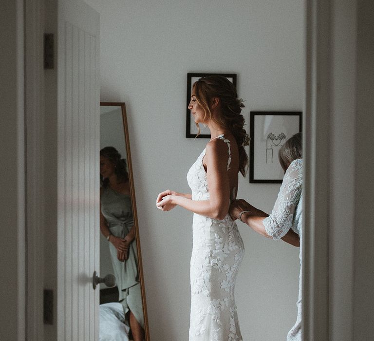 Bride wears floral embellished fitted Enzoani wedding dress and stands in front of the mirror on the morning of her wedding day