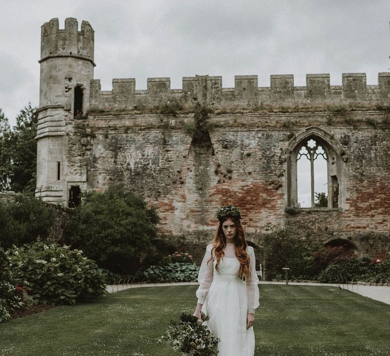 Bride in a wedding dress with sheer top layer and long sleeves wearing a foliage flower crown and holding a foliage bouquet standing in the gardens at Bishop's Palace, Wells