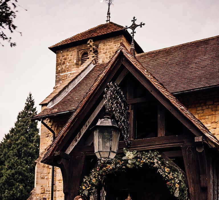 Bride in a long sleeve delicate lace wedding dress exits the church with the groom in grey trousers, a black morning suit jacket and yellow waistcoat