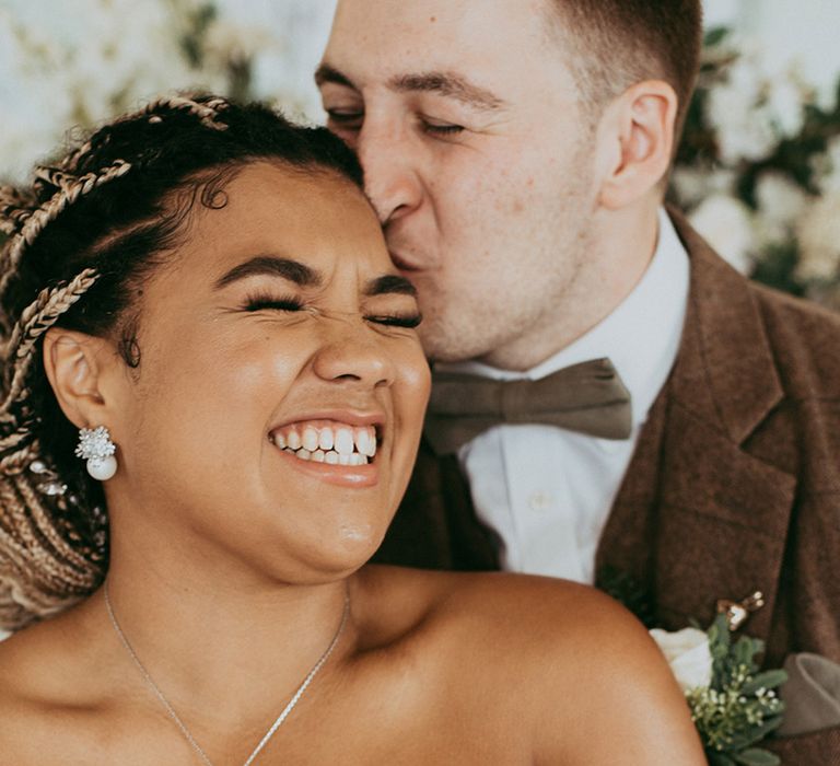 Bride smiling in a sweetheart off the shoulder wedding dress with a pearl and silver necklace and earrings with the groom in a brown suit