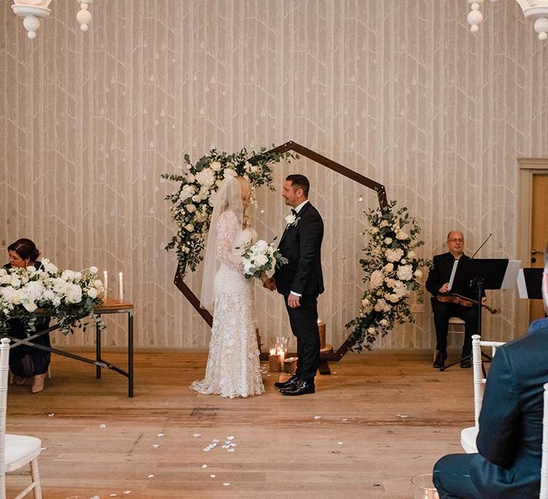 Bride & groom at Hampton Manor stand in front of hexagon arch complete with white floral decor 