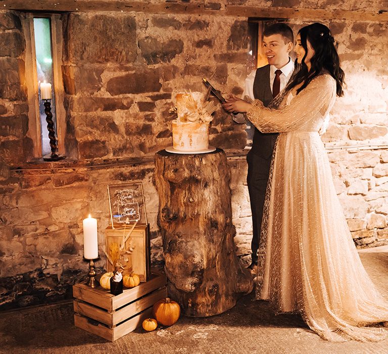 Bride & groom cut two-tier wedding cake on tree trunk in barn reception