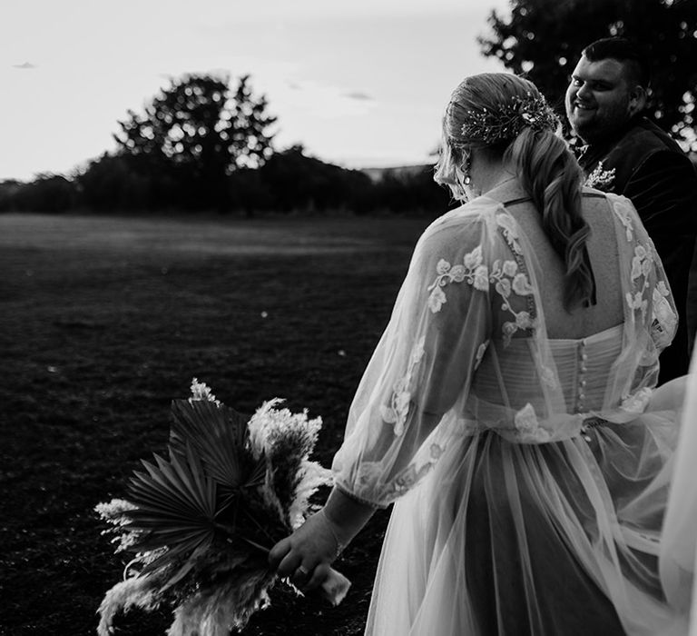 Bride in corset back detail wedding dress with with leaf detailed lace overlay, holding autumnal coloured bouquet with white pampas grass and an orange ribbon