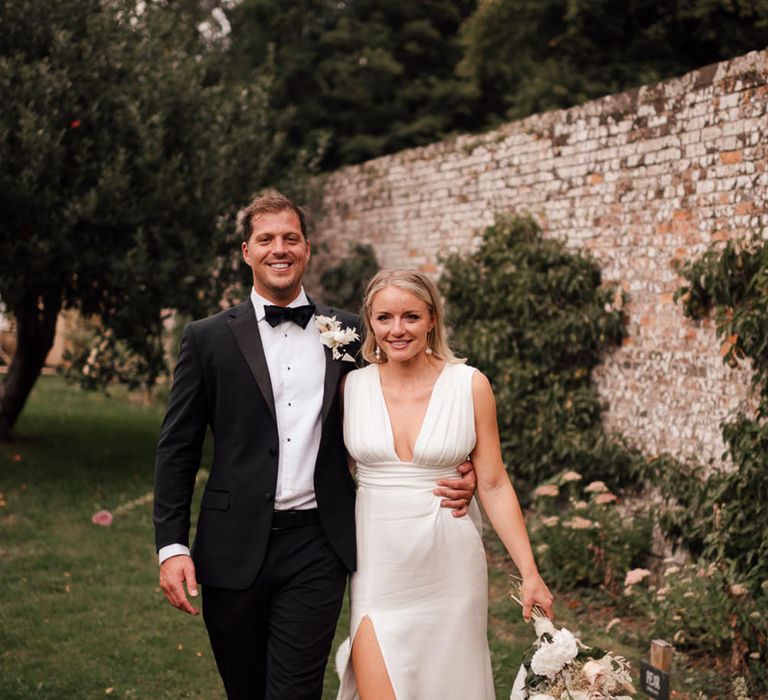 Groom in black tie with white flower buttonhole walking with the bride in a plunging satin wedding dress and orchid bouquet 