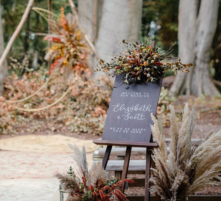 Hand made wood calligraphy welcome wedding sign on mahogany wooden easel with dried autumnal floral arrangements with white dried cotton stems, terra cotta celosia seeds and green foliage  