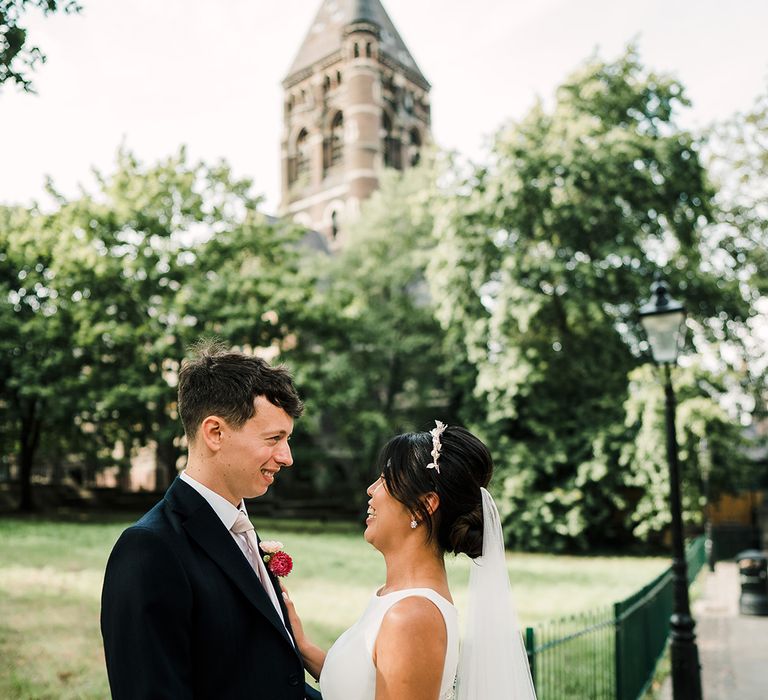 Bride in embellished crown and drop veil placed in her low up-do looks lovingly toward her groom during outdoor couples portraits