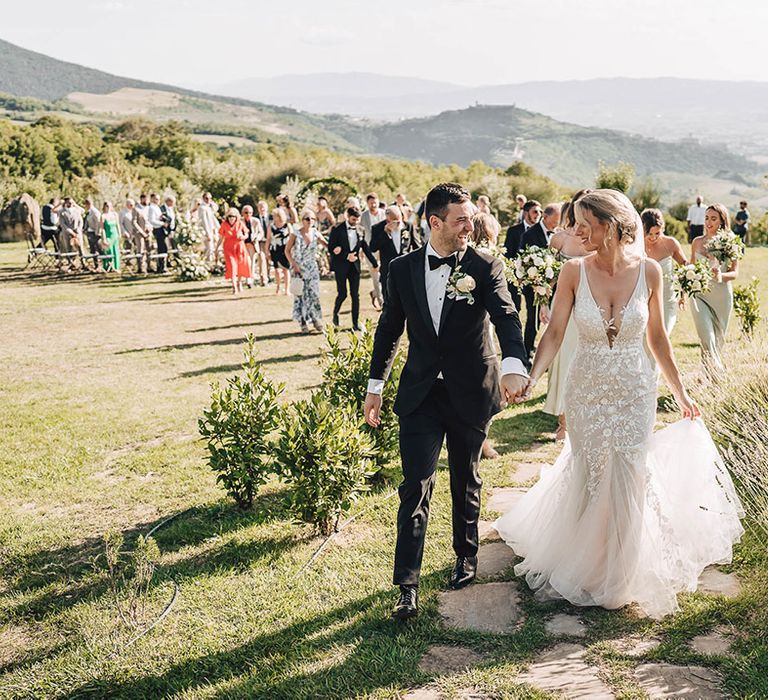 Bride wears lace plunge wedding dress with trumpet styled ruffled skirt and walks alongside her groom in black-tie 