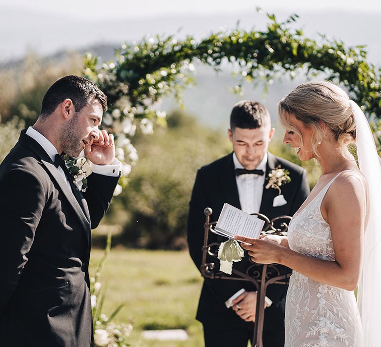 Bride in lace wedding dress reads personal wedding vows to emotional groom in black-tie