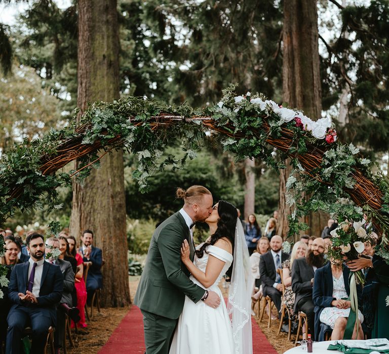 Groom in green suit kisses his bride in lace edge veil and off-the-shoulder wedding dress with sweetheart neckline 