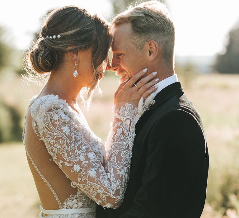 Bride in intricate lace wedding dress with long sleeves with an open back embracing the groom in black tie during golden hour 