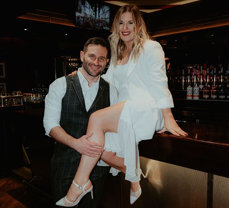 Bride in white ruffled wedding dress with pointed white shoes and blazer sits on the bar with the groom in checkered waistcoat 