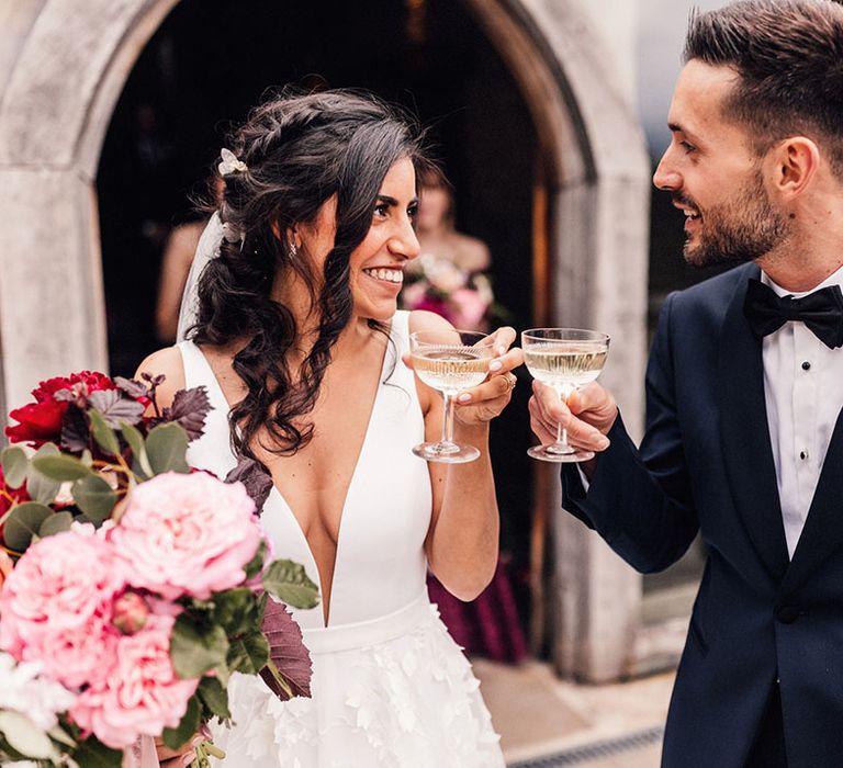 The groom in a traditional tuxedo shares a glass of champagne in coupe glasses with the bride after their wedding ceremony 