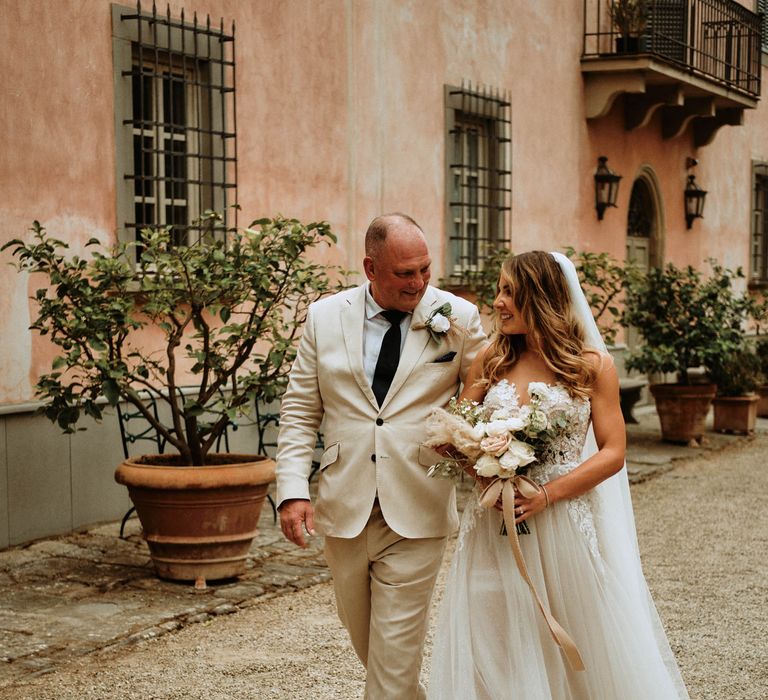 Father of the bride in linen suit walking with bride in embroidered tulle wedding dress and white rose bridal bouquet at Italian destination wedding