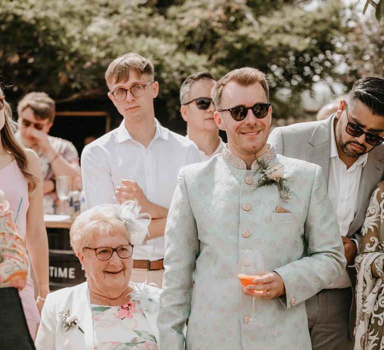 Groom in cream and gold Indian sherwani and white garden rose boutonniere holding hands with wedding guest in white dress with green and pink floral details 