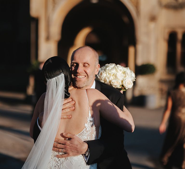 Bride in low scoop back lace wedding dress and church length veil holding white garden rose bridal bouquet hugging wedding guest in classic black tuxedo