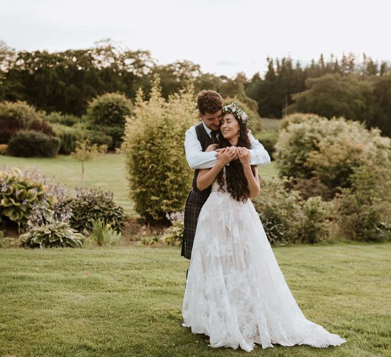 Groom in dark grey waistcoat and kilt embracing bride in 3d applique sheer overlay wedding dress with white garden rose flower crown and cathedral-length veil