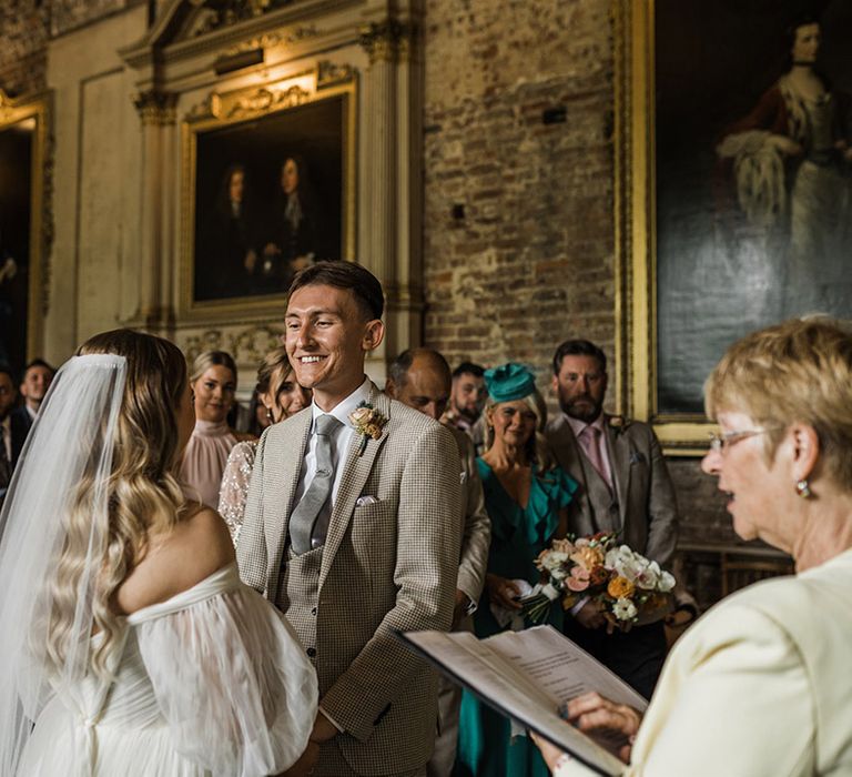 The groom in a three piece suit stands smiling and facing the bride for their wedding ceremony at St Giles House 