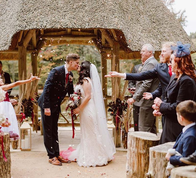 Groom in deep blue grooms blazer, grey waistcoat, red tie, red pocket square and mixed dried flower boutonniere kissing bride in sleeveless lace wedding dress and church length red ombre veil during colourful wedding confetti exit 