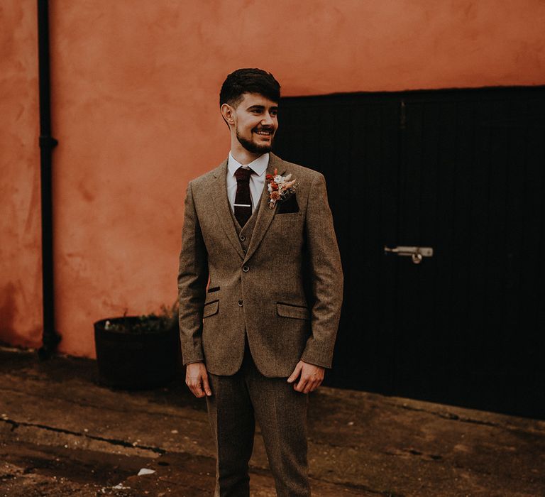 The groom wears a three piece grey suit with red tie and dried flower buttonhole for the rustic wedding 