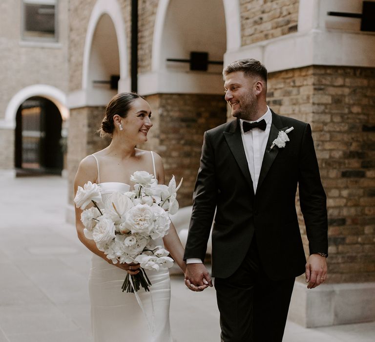 Bride carrying structural white wedding bouquet walking along with the groom in a black tux after their wedding 