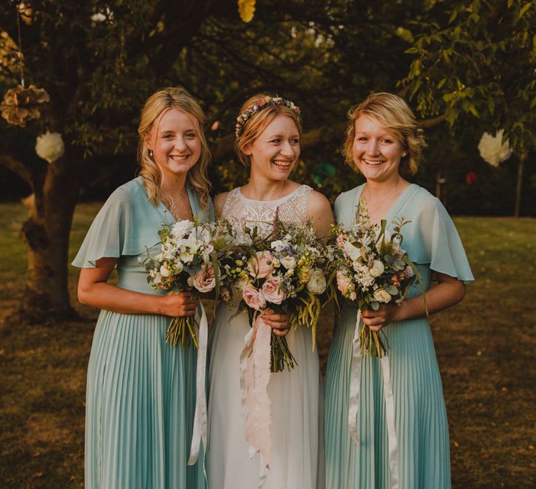 Bridesmaids in green pleated skirt midi dresses with bride in lace wedding dress and colourful flower crown 