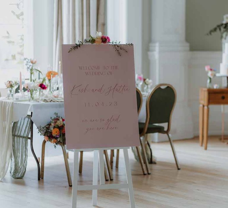 Pink classic wedding welcome sign on white easel with spring flowers on top at Freshman Hall wedding venue reception room