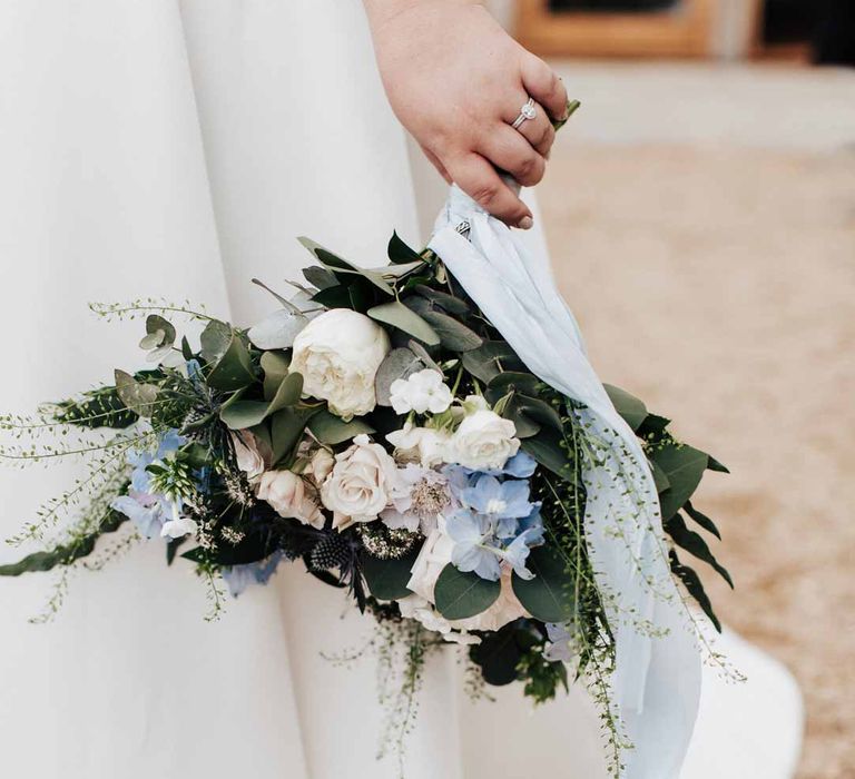 Bride holding white and blue eucalyptus, foliage, garden rose, gardenia, light blue delphinium and dried flower bridal bouquet tied with dusky blue ribbon 