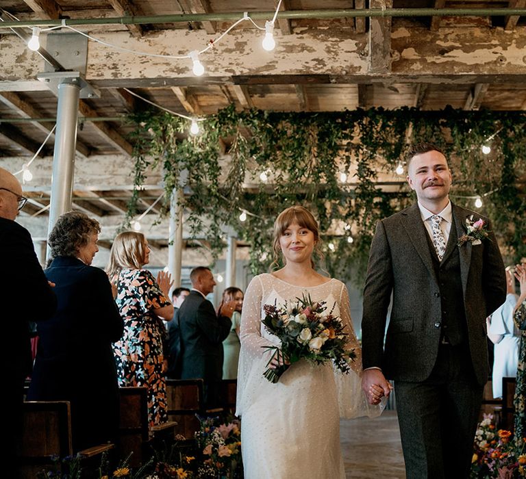 Groom in grey three piece suit with floral patterned tie walks back down the aisle with the bride in a dotted wedding dress 