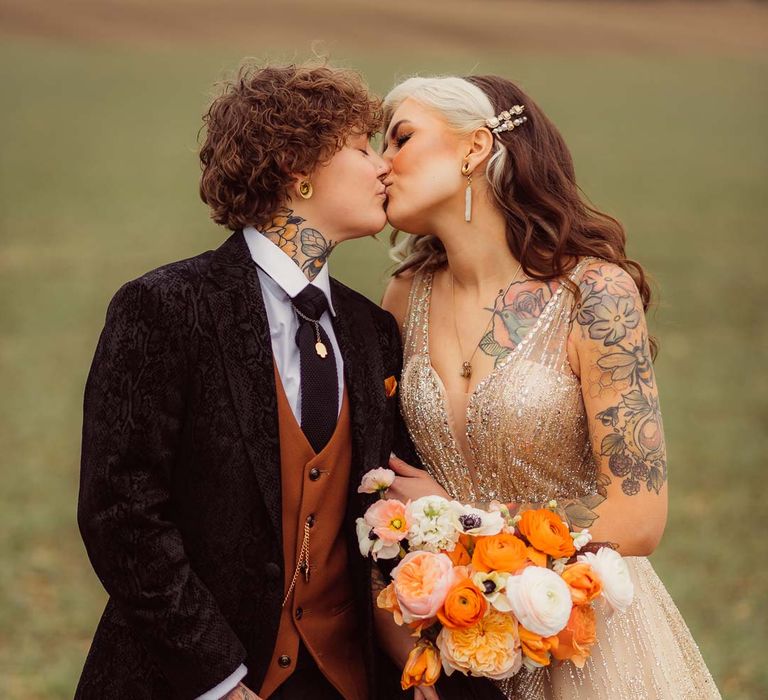 Bride in dark velvet bridal jacket with rust waistcoat and pocket square, black suit trousers and gold pocket watch standing with bride in sleeveless deep v-neck sparkly gold wedding dress holding orange garden rose, white poppy, light pink carnation and foliage bridal bouquet wrapped in sparkly gold ribbon at Kingscote Barn Tetbury wedding