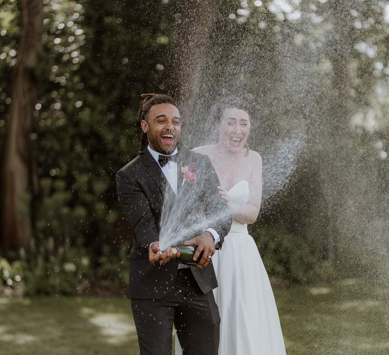 Groom in classic black tuxedo with bowtie and pink and orange wedding boutonniere doing wedding champagne pop photoshoot with bride in strapless sweetheart neckline wedding dress 