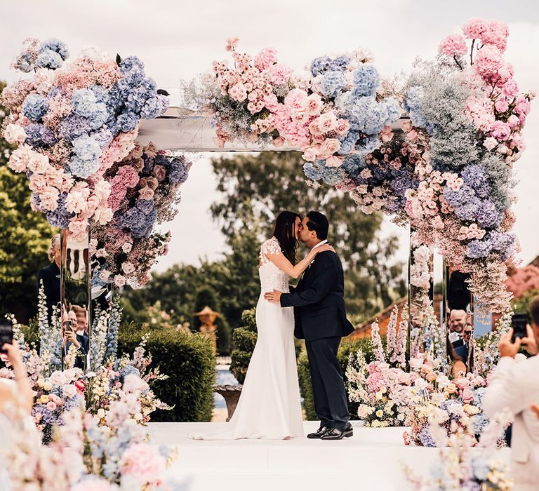 The bride and groom share their first kiss as a married couple at mirrored outdoor wedding ceremony set up 