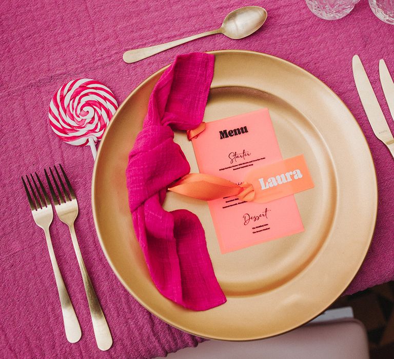 Lollipop wedding favours next to gold charger plates, coral place names and menus with pink napkins 