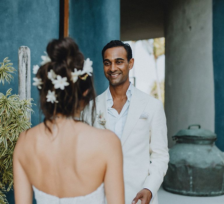 Smiling groom in white suit takes first look at bride in lace wedding dress and flower hair accessories