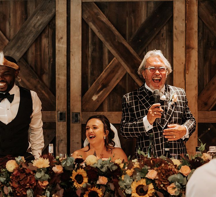 Groom in black tuxedo standing with the father of the groom in a black and white checkered suit reading out his speech with the bride seated in between