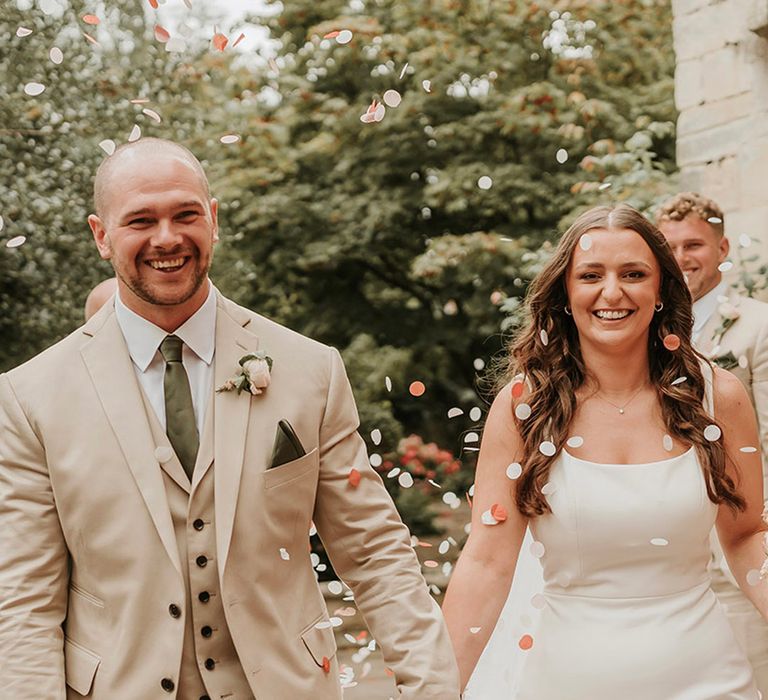 Colourful confetti moment for groom in three piece beige wedding suit with bride in satin slip wedding dress 