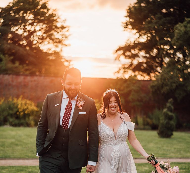 The groom in a dark green wedding suit walks with the bride in princess wedding dress during golden hour 