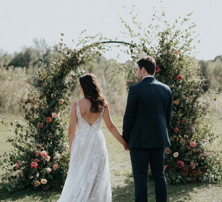 Bride and groom holding hands at outdoor wedding ceremony with floral moon gate 