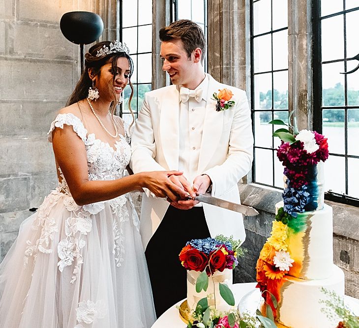 Bride in an off the shoulder wedding dress and groom in a white tuxedo jacket cutting the wedding cake with brightly coloured design 