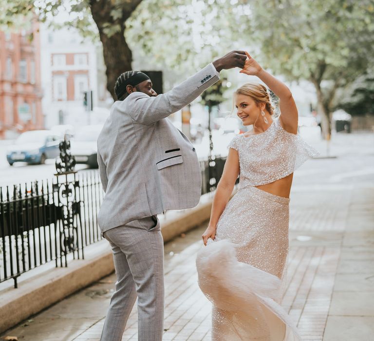 Groom in a light grey check suit twirling his bride in sequin bridal separates outside The Landmark London wedding venue 