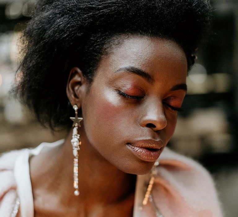 Wedding hairstyles for Black brides. A bride looks down to camera. She wears her afro hair swept back with neutral dewy make up.