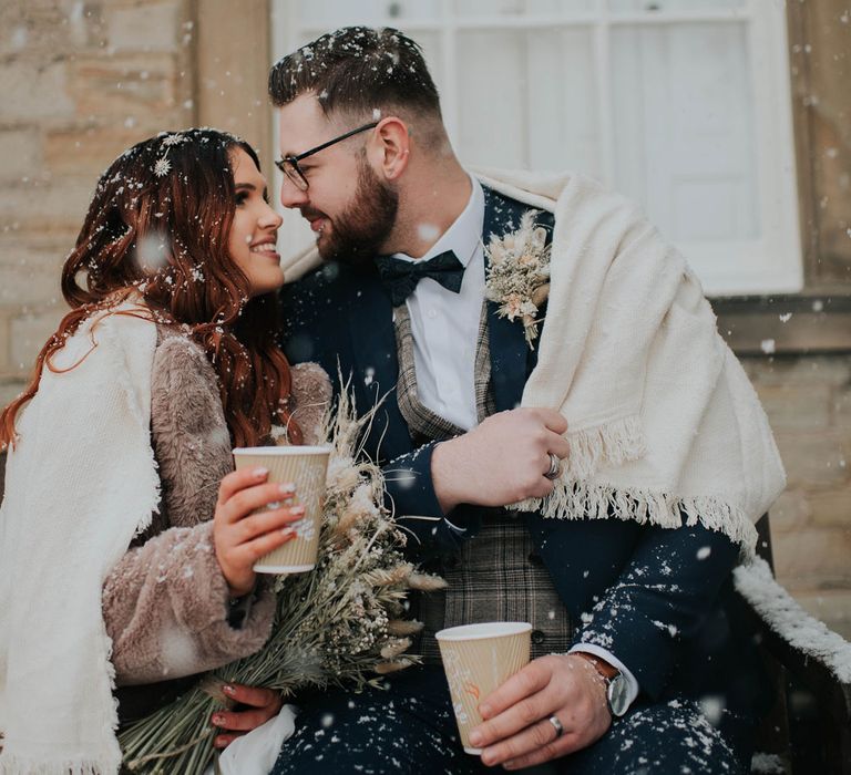 Bride in fur coat and wrap sits looking at groom in bow tie on steps during snowfall at Cannon Hall